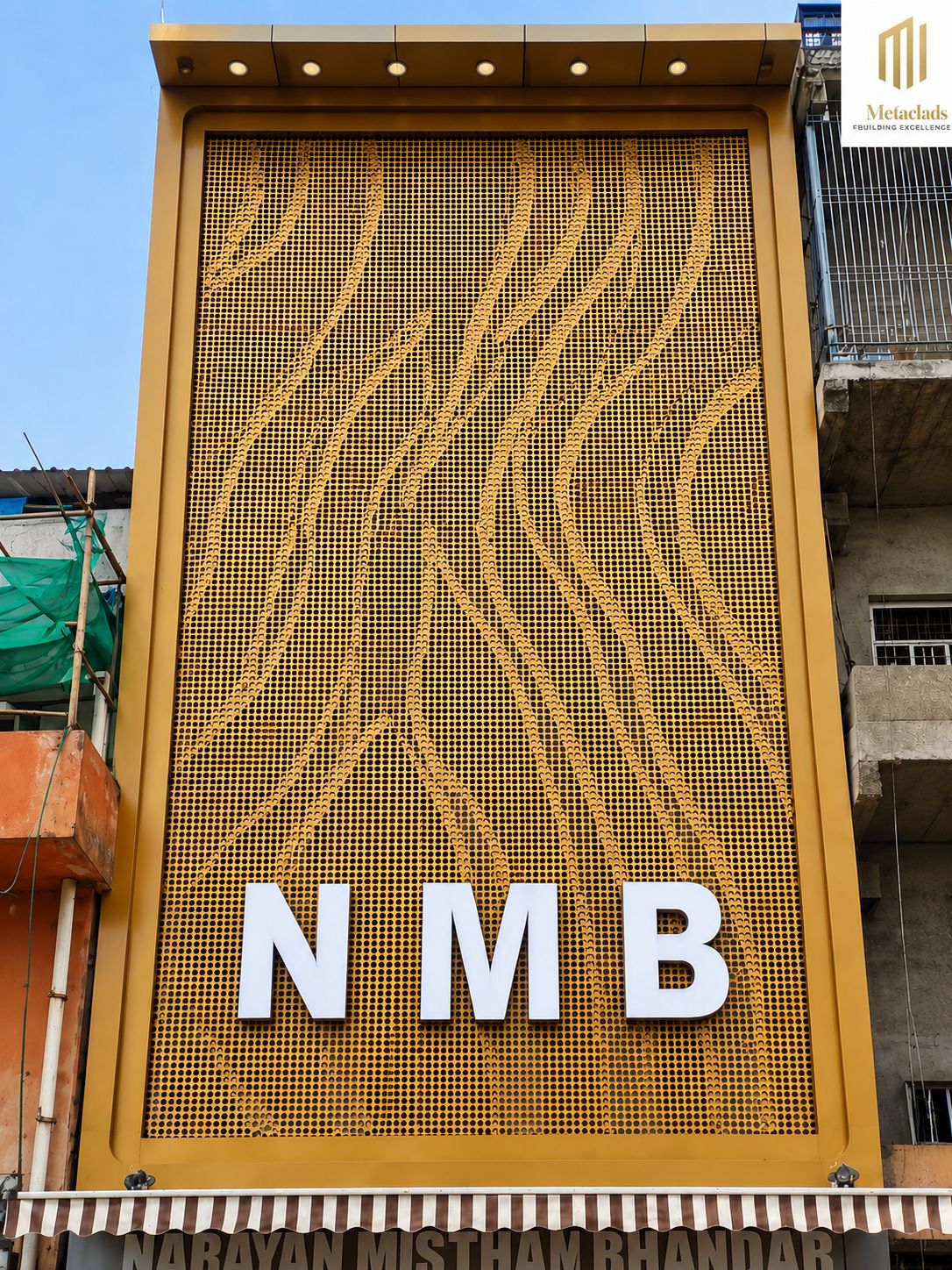 The NMB storefront in Agra: a full-height gold perforated aluminium screen whose dot pattern thins into flowing vertical striations, with white NMB lettering across the lower third and the Narayan Mistham Bhandar awning beneath. Scaffolding and a neighbouring unfinished block stand at either side.