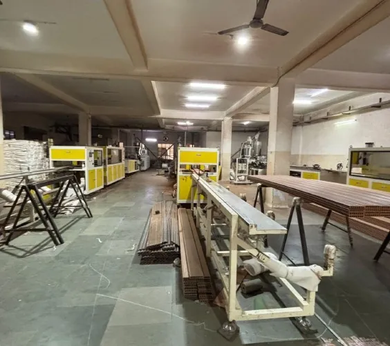 A workshop bay: yellow machines ranged along the far wall, steel trestles and a long roller conveyor running down the centre of the floor, lengths of profile stacked beside them, strip lights and a ceiling fan overhead.