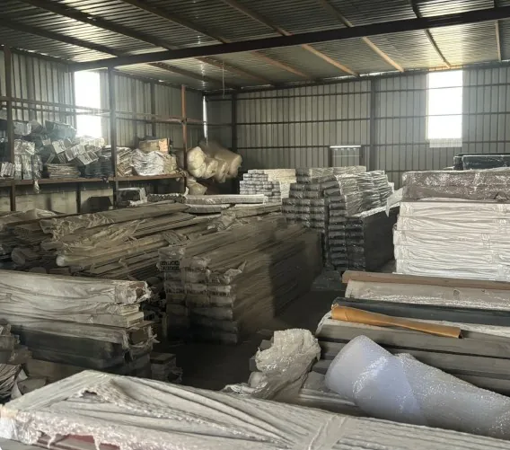 A storage bay stacked with film-wrapped panels and bundled boards on pallets, piled several courses high, under a corrugated roof with daylight coming through the roof lights.