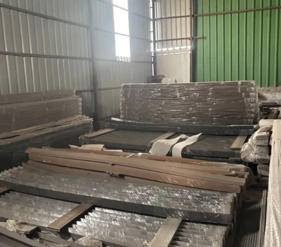 Bundles of dark boards and profiles racked in tall stacks against a green corrugated wall, a roof light above throwing light across the top of the pile.