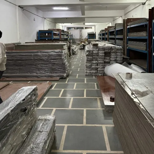 A storage aisle with wrapped panel stacks on blue steel racking down both sides, a tiled floor running away from the camera and rolls of sheet material at the far end.