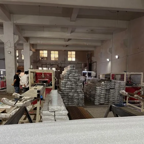 Two workers at a bench in a production hall, wrapped panel stacks piled behind them, a red machine to one side and lengths of decking laid across the foreground.