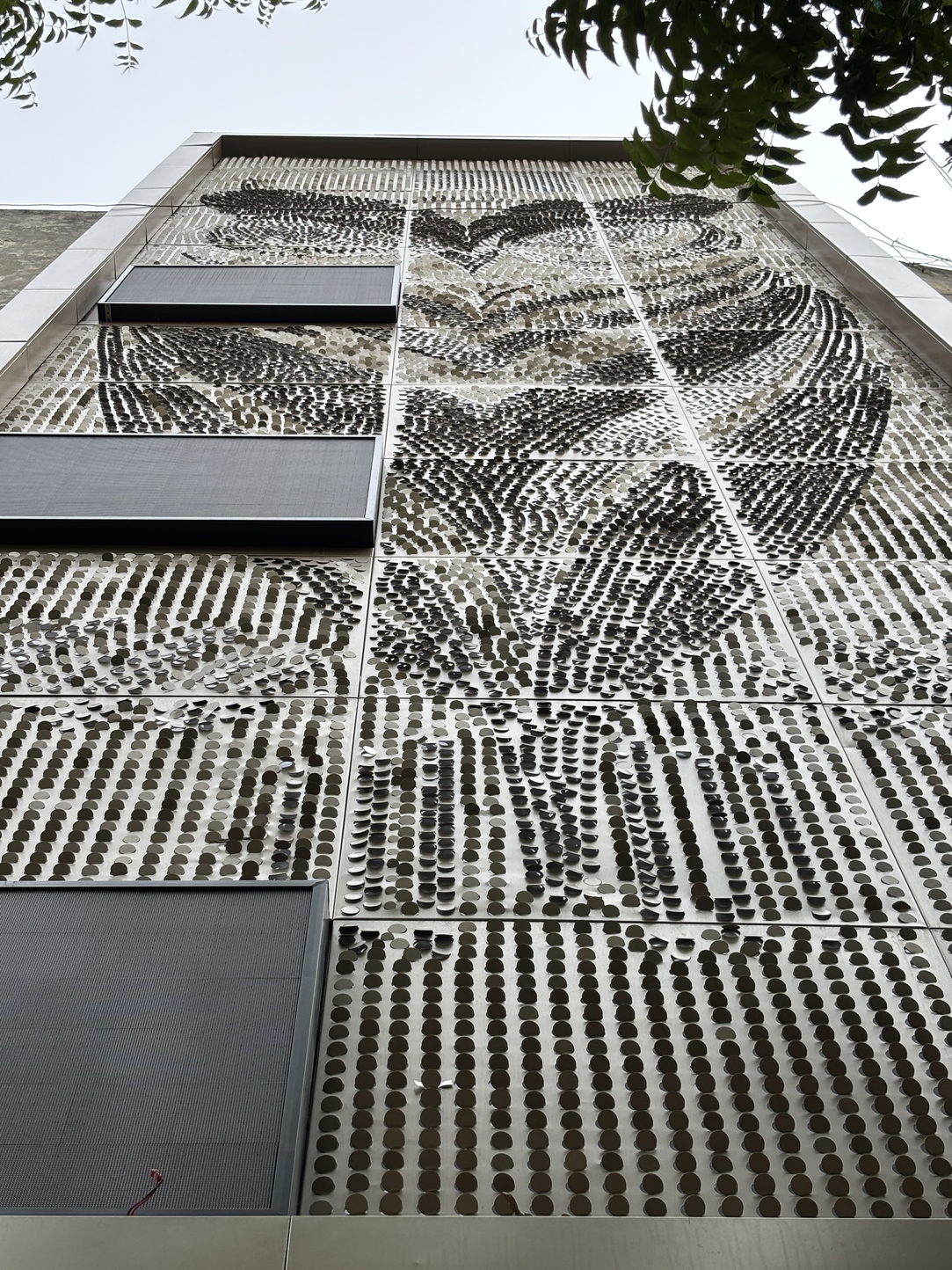 The Coin Matrix cladding close up, looking steeply up the face of the building. Small circular metal discs are fixed in rows across flat panels, each disc pivoted to its own angle, so the rows twist into dense swirling patterns high on the wall and settle into even lines lower down. The discs catch the light at different angles and the field reads as light and shadow rather than as a flat surface. Fine joints divide the cladding into rectangles, dark grey mesh louvre panels interrupt it on the left, and overcast sky and neem branches show across the top.