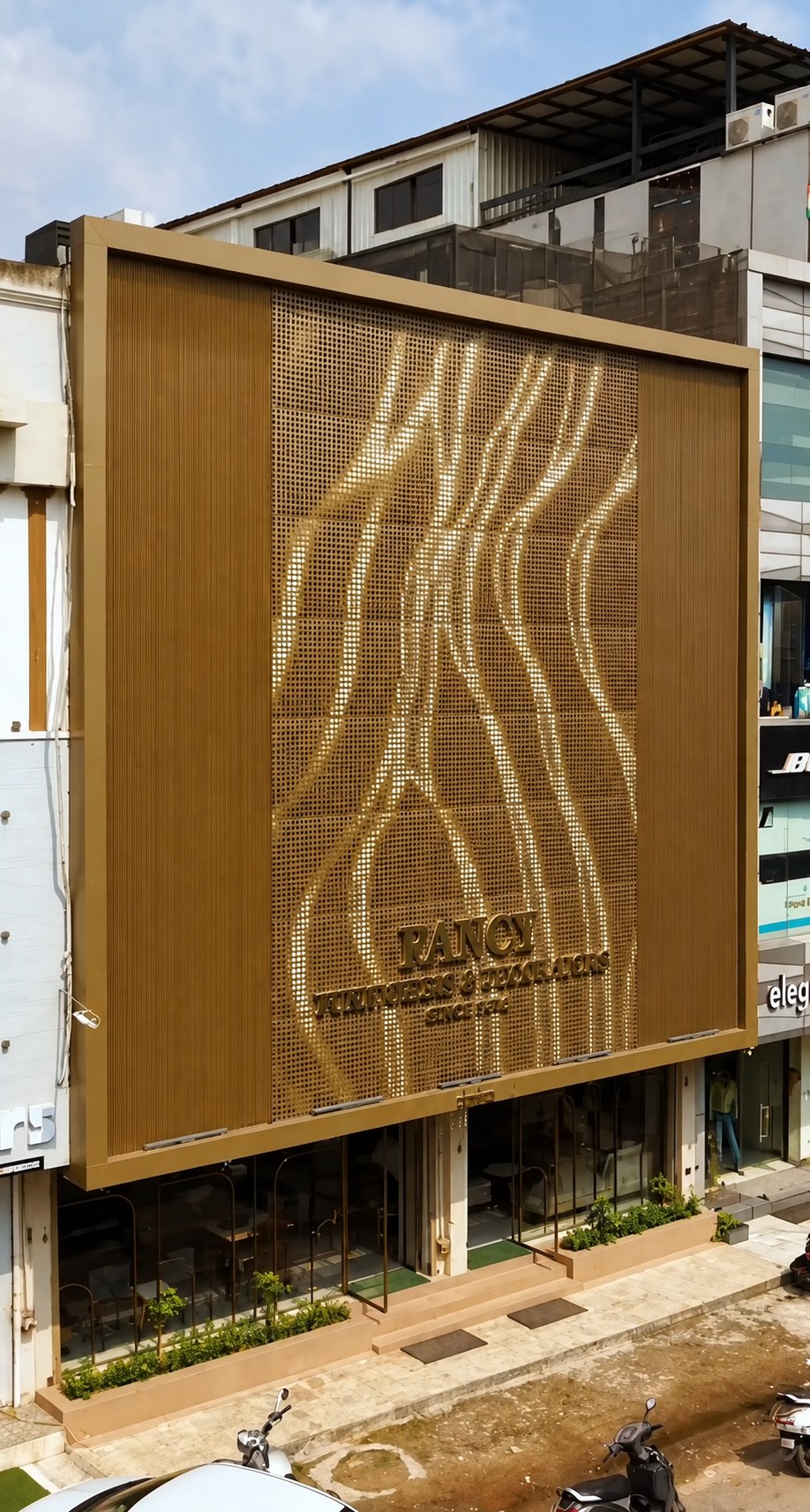 A retail elevation filled by a champagne-gold perforated screen in a heavy gold frame, its square holes graded so that the open runs curve down the sheet like hanging drapery and catch the light. Fluted vertical panels in the same gold close the elevation on both sides. Raised lettering reading FANCY FURNISHERS & DECORATORS sits low on the screen, and the glazed retail floor below shows furniture and planting, with scooters parked at the kerb.