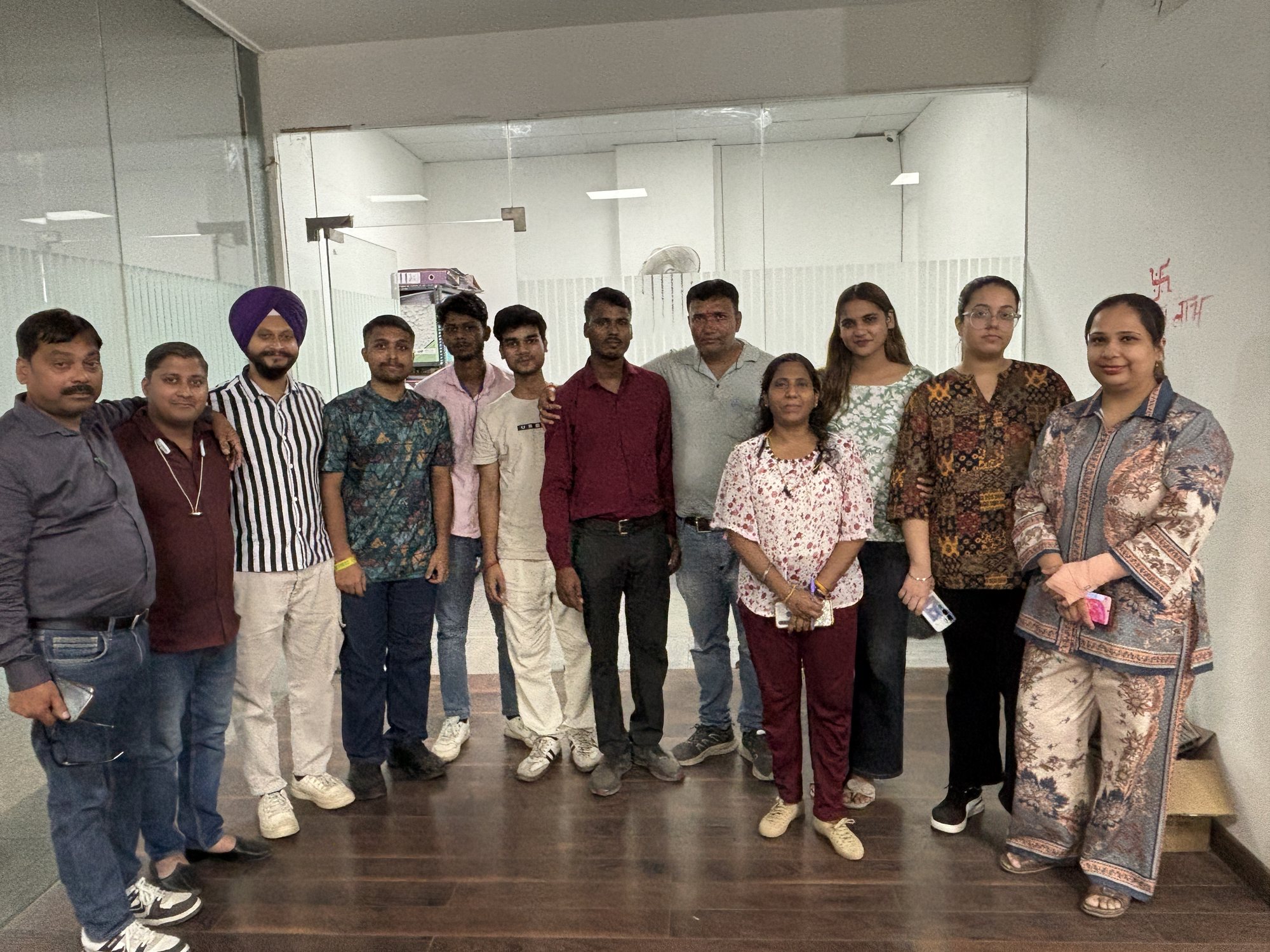 Twelve members of the head office team standing together for a group photograph in a glass-partitioned office with a dark wood floor, files and a desk visible through the partition behind them.