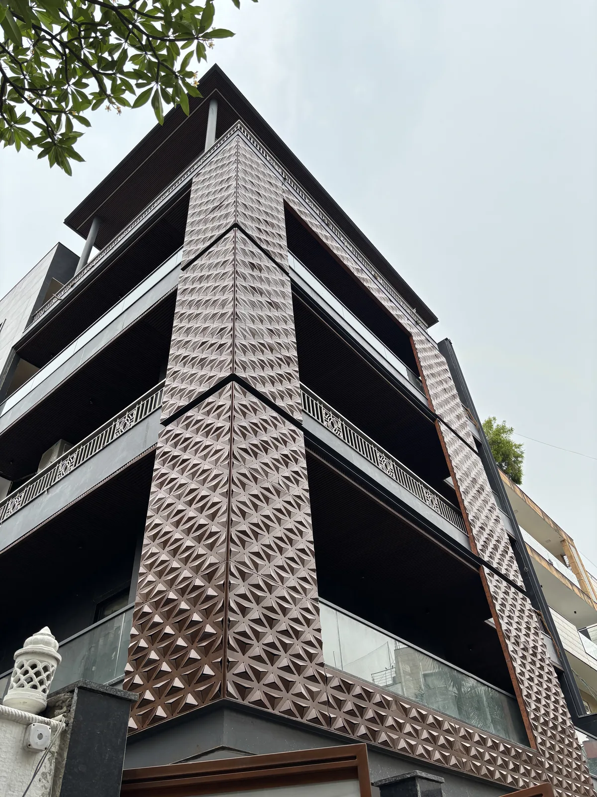 A triangular cassette façade turning the corner of a four-storey building, seen looking up from the street. The cassettes are pressed into three-sided pyramids that run in a continuous field up the corner column and across the returns on either side, each facet catching the overcast light at its own angle so the surface reads as a shifting pattern of pale and shadowed triangles rather than a flat skin. The metal is a warm copper tone. Recessed balconies with dark soffits and slim glass balustrades step back on the right, and a frangipani branch crosses the top-left corner.