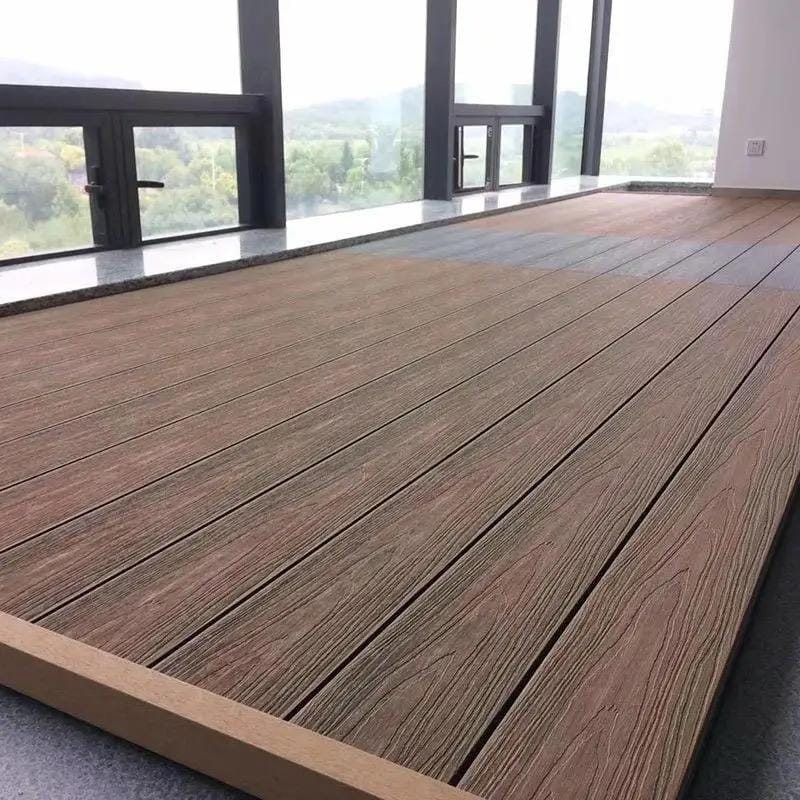 A raised deck of wide walnut-toned composite boards, laid in long parallel runs with a narrow shadow gap between each, finished with a square edge trim. Floor-to-ceiling glazing behind it looks out onto wooded hills.