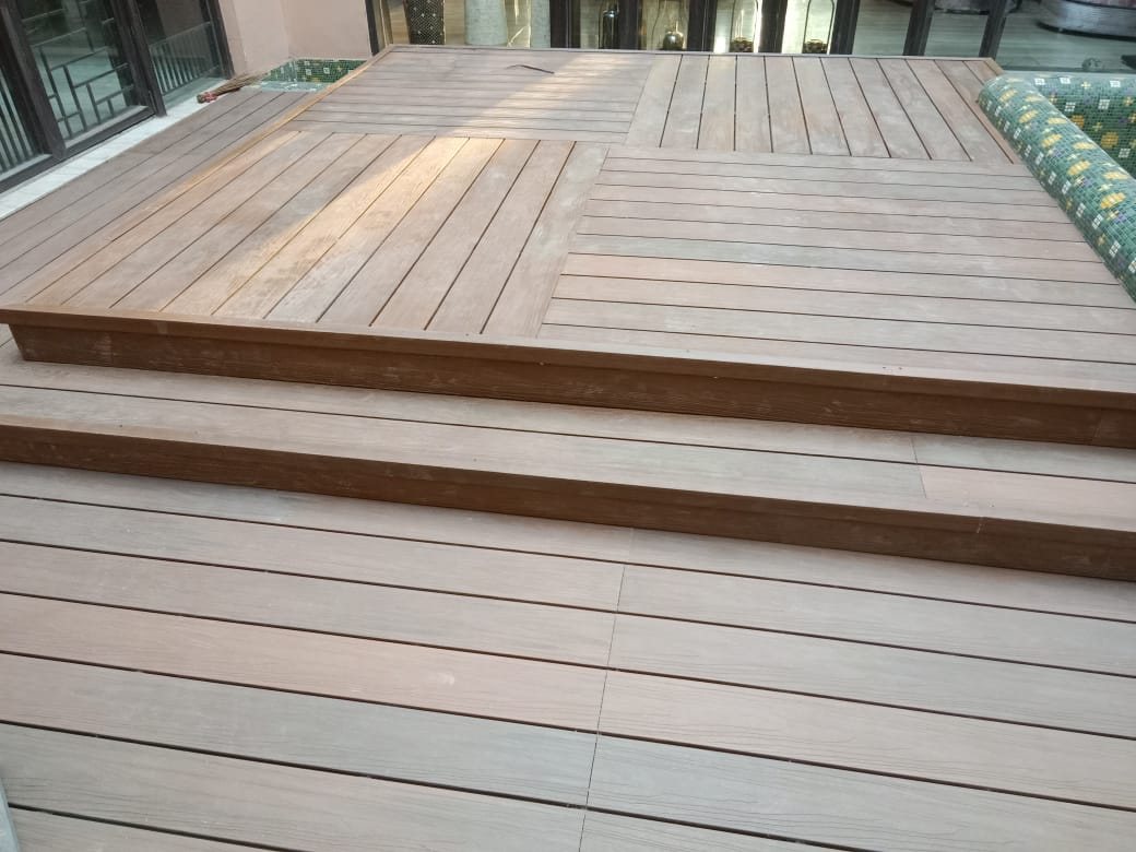 A terrace decked in pale ash-grey composite boards, stepping down in two broad levels with a darker board closing the nose of each step. Mosaic-tiled pool coping runs along the far right edge.