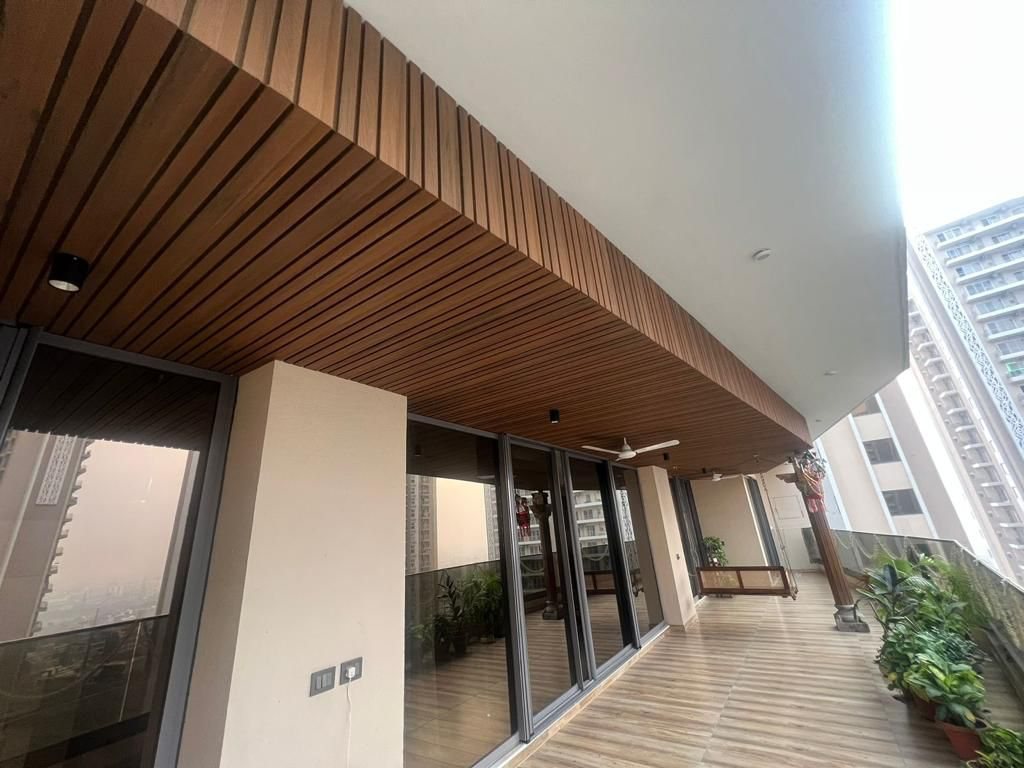 A long balcony with its soffit lined in closely ranked composite battens in a warm timber tone, running the length of the overhang and turning down its outer face. Sliding glazing runs along the left, planters along the right, and a city skyline shows beyond.