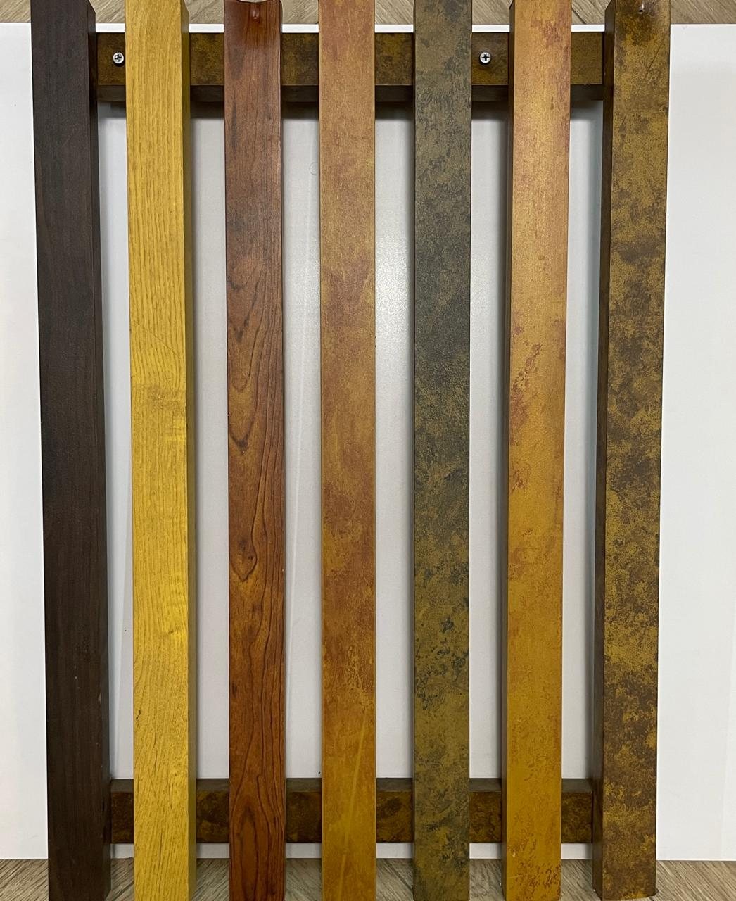 Seven composite rafter sections fixed vertically across a white sample board, each in a different finish — dark walnut, gold, red timber grain, rust, mossy green-brown and weathered ochre — so the range can be compared side by side.